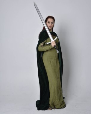 Fantezi kadife pelerinli yeşil ortaçağ elbisesi giyen kızıl saçlı kızın tam boy portresi. Stüdyonun arka planında yalnız poz vermek.