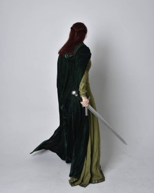 Fantezi kadife pelerinli yeşil ortaçağ elbisesi giyen kızıl saçlı kızın tam boy portresi. Stüdyonun arka planında yalnız poz vermek.