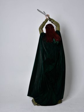 Fantezi kadife pelerinli yeşil ortaçağ elbisesi giyen kızıl saçlı kızın tam boy portresi. Stüdyonun arka planında yalnız poz vermek.