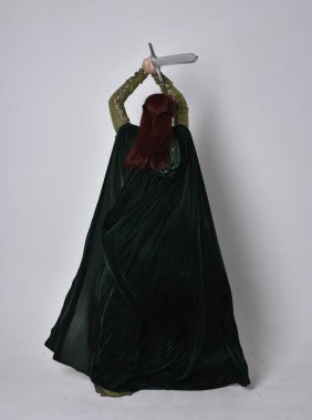 Fantezi kadife pelerinli yeşil ortaçağ elbisesi giyen kızıl saçlı kızın tam boy portresi. Stüdyonun arka planında yalnız poz vermek.