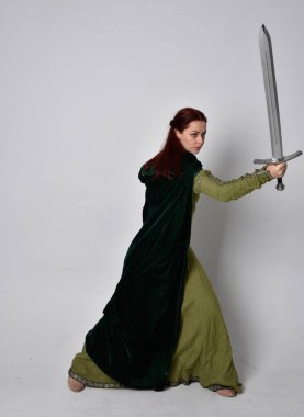 Fantezi kadife pelerinli yeşil ortaçağ elbisesi giyen kızıl saçlı kızın tam boy portresi. Stüdyonun arka planında yalnız poz vermek.