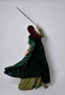 Fantezi kadife pelerinli yeşil ortaçağ elbisesi giyen kızıl saçlı kızın tam boy portresi. Stüdyonun arka planında yalnız poz vermek.