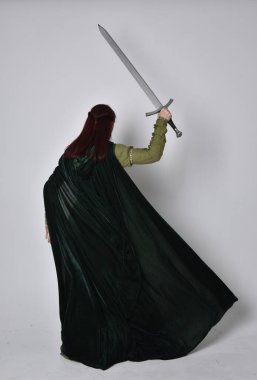 Fantezi kadife pelerinli yeşil ortaçağ elbisesi giyen kızıl saçlı kızın tam boy portresi. Stüdyonun arka planında yalnız poz vermek.