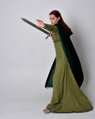 Fantezi kadife pelerinli yeşil ortaçağ elbisesi giyen kızıl saçlı kızın tam boy portresi. Stüdyonun arka planında yalnız poz vermek.