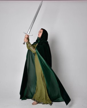 Fantezi kadife pelerinli yeşil ortaçağ elbisesi giyen kızıl saçlı kızın tam boy portresi. Stüdyonun arka planında yalnız poz vermek.