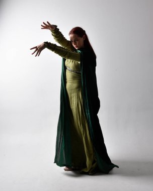 Kızıl saçlı, yeşil, gölgeli arka ışıklandırmalı ortaçağ elbisesi giyen bir kızın tam boy portresi. Stüdyonun arka planında yalnız poz vermek.