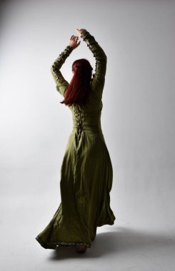 Kızıl saçlı, yeşil, gölgeli arka ışıklandırmalı ortaçağ elbisesi giyen bir kızın tam boy portresi. Stüdyonun arka planında yalnız poz vermek.