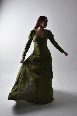 Kızıl saçlı, yeşil, gölgeli arka ışıklandırmalı ortaçağ elbisesi giyen bir kızın tam boy portresi. Stüdyonun arka planında yalnız poz vermek.