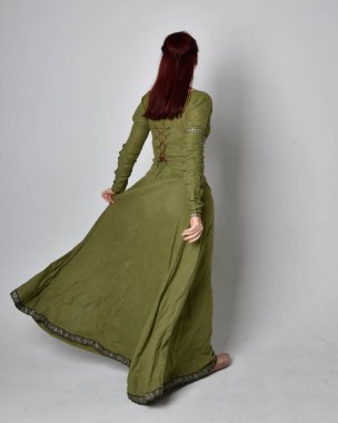 Yeşil ortaçağ elbisesi giyen kızıl saçlı kızın tam boy portresi. Stüdyonun arka planında yalnız poz vermek.