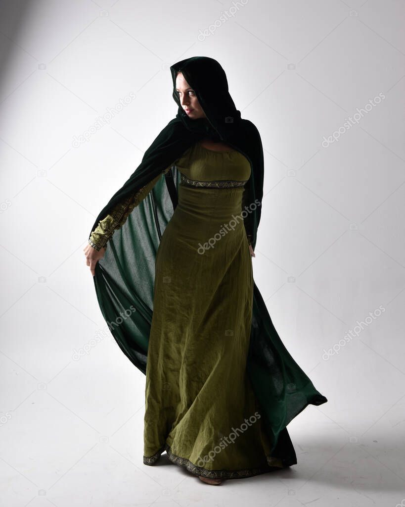 full length portrait of red haired girl wearing celtic, green medieval gown with shadowy backlighting. Standing pose isolated against a studio background.