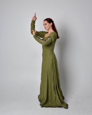 Yeşil ortaçağ elbisesi giyen kızıl saçlı kızın tam boy portresi. Stüdyonun arka planında yalnız poz vermek.