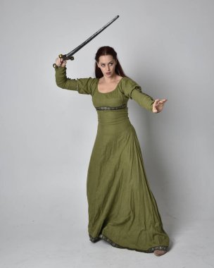 Yeşil ortaçağ elbisesi giyen kızıl saçlı kızın tam boy portresi. Duruş, kılıç tutma, stüdyo arka planında izole edilmiş..