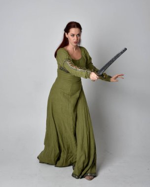 Yeşil ortaçağ elbisesi giyen kızıl saçlı kızın tam boy portresi. Duruş, kılıç tutma, stüdyo arka planında izole edilmiş..