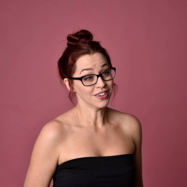 Pembe stüdyo arka planında gözlüklü, etkileyici yüz hatları olan kızıl saçlı bir kadının portresi..