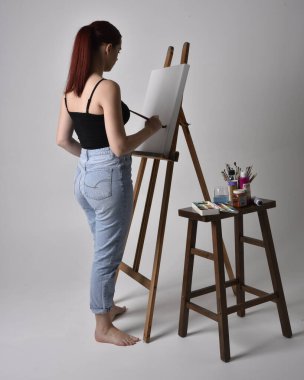 Normal kot pantolon ve kolsuz bluz giyen kızıl saçlı bir kızın tam boy portresi. Stüdyonun arka planına karşı sehpada bir tuval boyuyor..