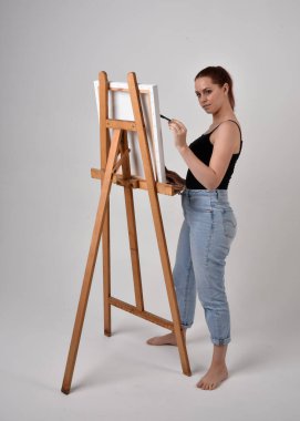 Normal kot pantolon ve kolsuz bluz giyen kızıl saçlı bir kızın tam boy portresi. Stüdyonun arka planına karşı sehpada bir tuval boyuyor..