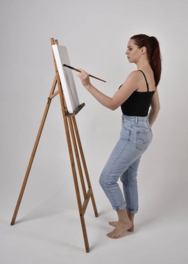 Normal kot pantolon ve kolsuz bluz giyen kızıl saçlı bir kızın tam boy portresi. Stüdyonun arka planına karşı sehpada bir tuval boyuyor..