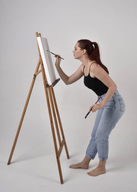 Normal kot pantolon ve kolsuz bluz giyen kızıl saçlı bir kızın tam boy portresi. Stüdyonun arka planına karşı sehpada bir tuval boyuyor..