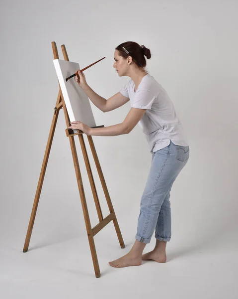 Full length portrait of a red haired artist girl wearing casual jeans and white shirt.  standing pose painting a canvas on an easel, against a studio background.