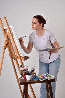 Sıradan kıyafetler giyen kızıl saçlı bir sanatçının portresi. Stüdyonun arka planına karşı sehpada bir tuval boyuyor..