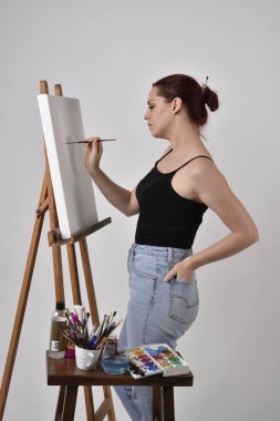 Sıradan kıyafetler giyen kızıl saçlı bir sanatçının portresi. Stüdyonun arka planına karşı sehpada bir tuval boyuyor..