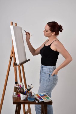 Sıradan kıyafetler giyen kızıl saçlı bir sanatçının portresi. Stüdyonun arka planına karşı sehpada bir tuval boyuyor..