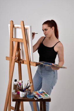 Sıradan kıyafetler giyen kızıl saçlı bir sanatçının portresi. Stüdyonun arka planına karşı sehpada bir tuval boyuyor..