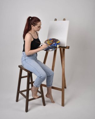 Normal kot pantolon ve beyaz gömlek giyen kızıl saçlı bir kızın tam boy portresi. Sandalyeye oturmak, bir sehpaya resim yapmak stüdyo arka planına karşı.