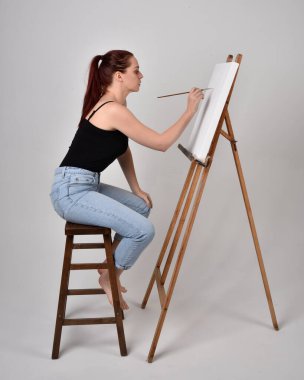 Normal kot pantolon ve beyaz gömlek giyen kızıl saçlı bir kızın tam boy portresi. Sandalyeye oturmak, bir sehpaya resim yapmak stüdyo arka planına karşı.