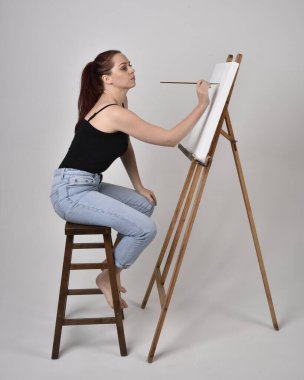 Normal kot pantolon ve beyaz gömlek giyen kızıl saçlı bir kızın tam boy portresi. Sandalyeye oturmak, bir sehpaya resim yapmak stüdyo arka planına karşı.