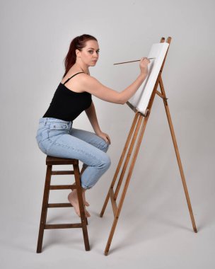 Normal kot pantolon ve beyaz gömlek giyen kızıl saçlı bir kızın tam boy portresi. Sandalyeye oturmak, bir sehpaya resim yapmak stüdyo arka planına karşı.