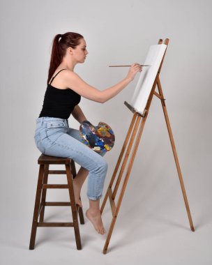 Normal kot pantolon ve beyaz gömlek giyen kızıl saçlı bir kızın tam boy portresi. Sandalyeye oturmak, bir sehpaya resim yapmak stüdyo arka planına karşı.