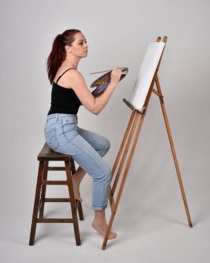 Normal kot pantolon ve beyaz gömlek giyen kızıl saçlı bir kızın tam boy portresi. Sandalyeye oturmak, bir sehpaya resim yapmak stüdyo arka planına karşı.