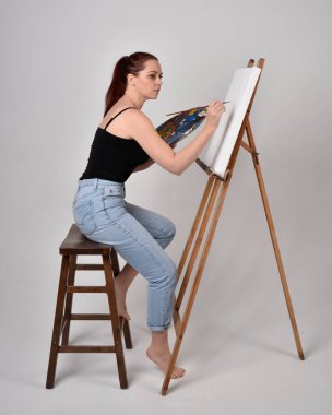 Normal kot pantolon ve beyaz gömlek giyen kızıl saçlı bir kızın tam boy portresi. Sandalyeye oturmak, bir sehpaya resim yapmak stüdyo arka planına karşı.