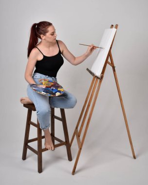 Normal kot pantolon ve beyaz gömlek giyen kızıl saçlı bir kızın tam boy portresi. Sandalyeye oturmak, bir sehpaya resim yapmak stüdyo arka planına karşı.