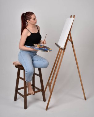 Normal kot pantolon ve beyaz gömlek giyen kızıl saçlı bir kızın tam boy portresi. Sandalyeye oturmak, bir sehpaya resim yapmak stüdyo arka planına karşı.