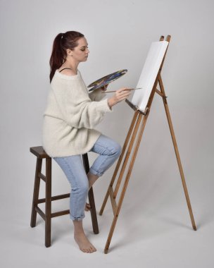 Normal kot pantolon ve beyaz gömlek giyen kızıl saçlı bir kızın tam boy portresi. Sandalyeye oturmak, bir sehpaya resim yapmak stüdyo arka planına karşı.