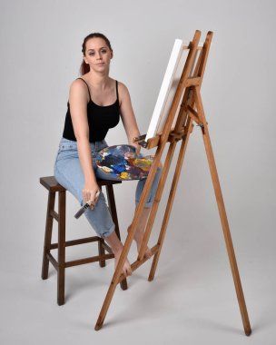 Normal kot pantolon ve beyaz gömlek giyen kızıl saçlı bir kızın tam boy portresi. Sandalyeye oturmak, bir sehpaya resim yapmak stüdyo arka planına karşı.