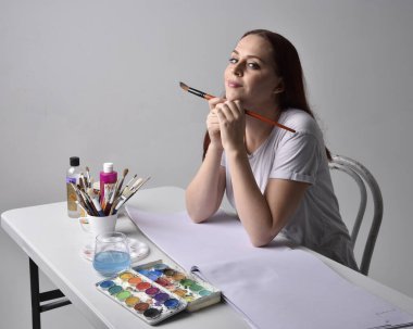 Beyaz gömlek giyen kızıl saçlı bir ressamın yakın portresi. Stüdyo arka planında suluboya resimlerle bir masa başında oturmak..