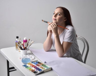 Beyaz gömlek giyen kızıl saçlı bir ressamın yakın portresi. Stüdyo arka planında suluboya resimlerle bir masa başında oturmak..