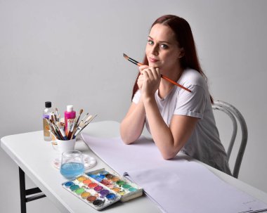 Beyaz gömlek giyen kızıl saçlı bir ressamın yakın portresi. Stüdyo arka planında suluboya resimlerle bir masa başında oturmak..