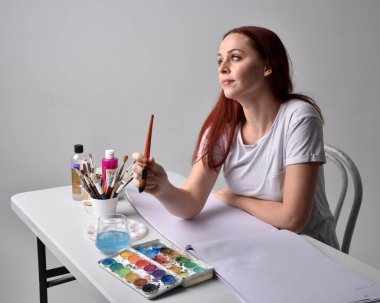 Beyaz gömlek giyen kızıl saçlı bir ressamın yakın portresi. Stüdyo arka planında suluboya resimlerle bir masa başında oturmak..