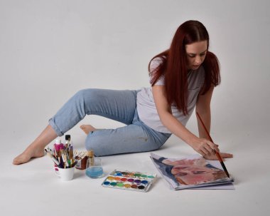 Beyaz gömlek giyen kızıl saçlı bir ressamın yakın portresi. Stüdyo arka planında suluboya boyalarla yerde oturuyor..