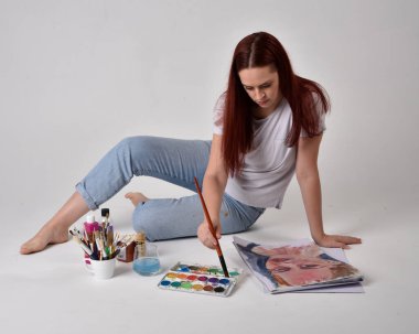 Beyaz gömlek giyen kızıl saçlı bir ressamın yakın portresi. Stüdyo arka planında suluboya boyalarla yerde oturuyor..