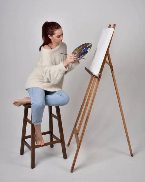 Full length portrait of a red haired artist girl wearing casual jeans and white shirt.  Sitting pose on chair, painting a canvas on an easel against a studio background