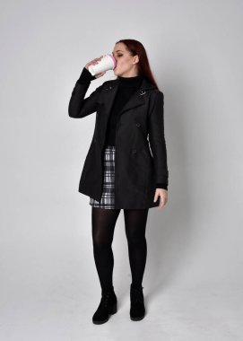 Siyah ceket, tartan etek ve çizme giyen kızıl saçlı bir kızın tam boy portresi. Stüdyo geçmişine karşı poz vermek.