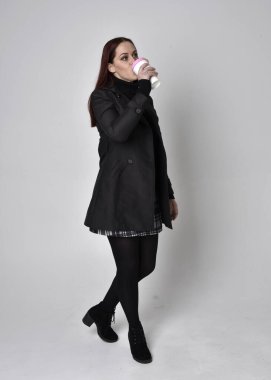 Siyah ceket, tartan etek ve çizme giyen kızıl saçlı bir kızın tam boy portresi. Stüdyo geçmişine karşı poz vermek.