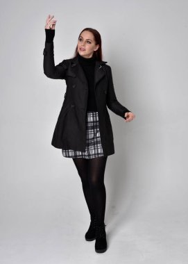 Siyah ceket, tartan etek ve çizme giyen kızıl saçlı bir kızın tam boy portresi. Stüdyo geçmişine karşı poz vermek.