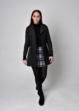 Siyah ceket, tartan etek ve çizme giyen kızıl saçlı bir kızın tam boy portresi. Stüdyo geçmişine karşı poz vermek.
