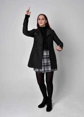 Siyah ceket, tartan etek ve çizme giyen kızıl saçlı bir kızın tam boy portresi. Stüdyo geçmişine karşı poz vermek.
